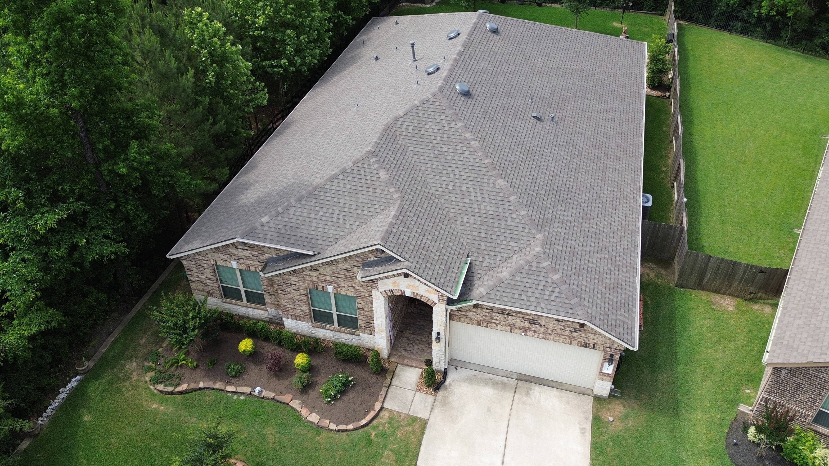 Aerial drone view of a single-story brick home with a newly installed light gray architectural shingle roof on a wooded lot