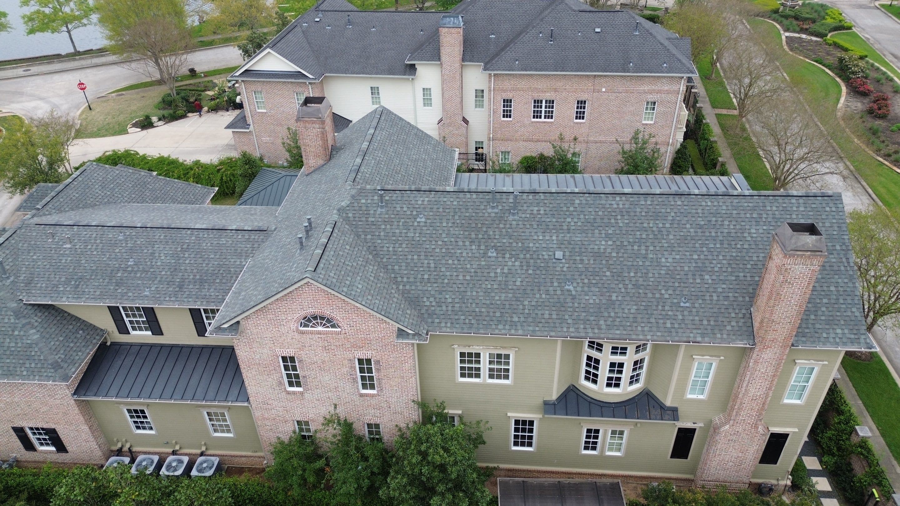 Drone view of a large brick estate home with new gray architectural shingles and standing-seam metal accent roofs